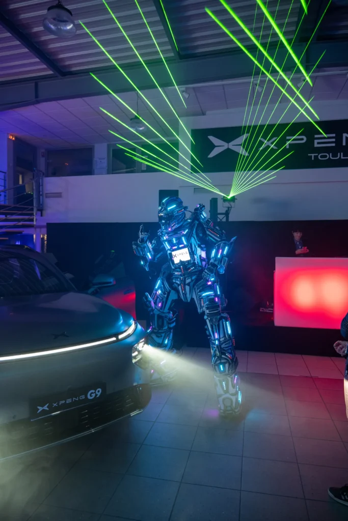 Performeur en costume lumineux devant un véhicule, sous un réseau de faisceaux laser verts