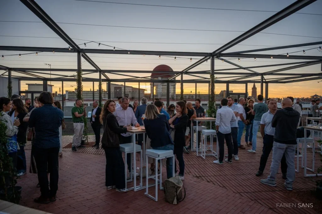Invités autour de mange-debout sur un toit-terrasse de Toulouse au coucher du soleil, guirlandes tendues sous l’ossature métallique